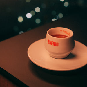 A small white ceramic espresso cup with the brand name RUFINO printed in red, sitting on a matching saucer. The cup is filled with a espresso and is placed on a dark table with a blurred dark background featuring soft bokeh lights.